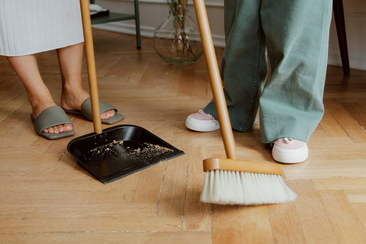 hero-img-01 Close-up of people cleaning wooden floor with broom and dustpan indoors.